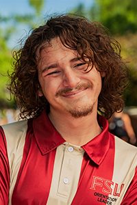 male student smiling, entring the frame from the left at an angle, with mid-length brown curly hair and a moustache and goatee