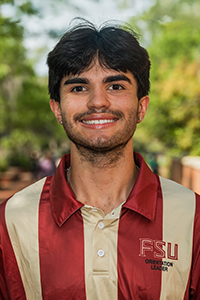 smiling male student with short brown hair parted in the middle and a bushy moustache and thinner beard