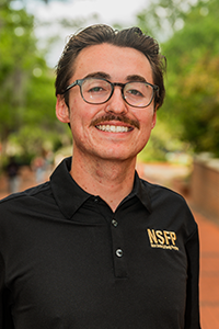 smiling male student with short brown hair parted in the middle, with a moustache and thick black glasses