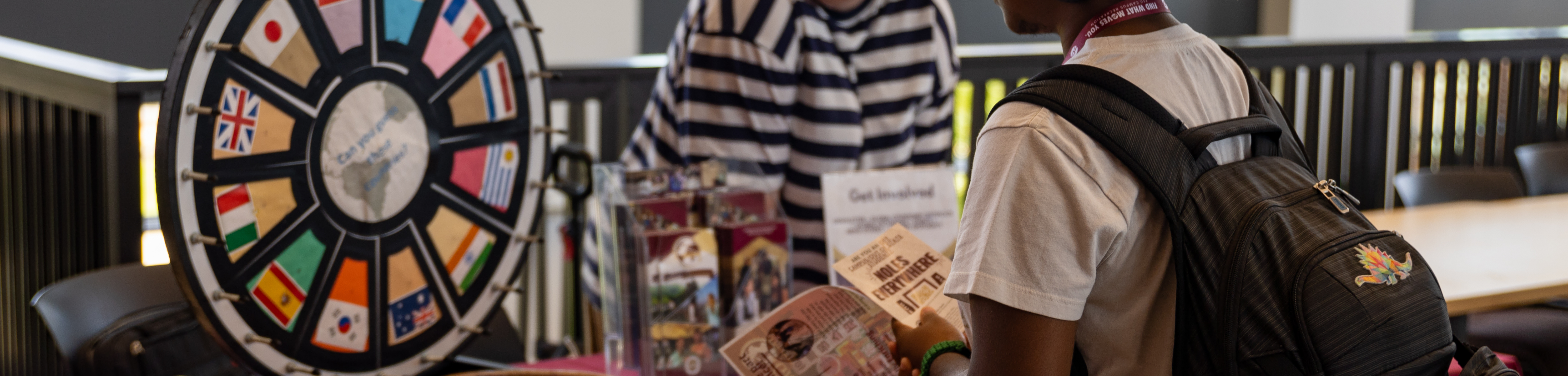 A student with a backpack looks at a pamphlet while standing at a table with a colorful prize wheel decorated with international flags.