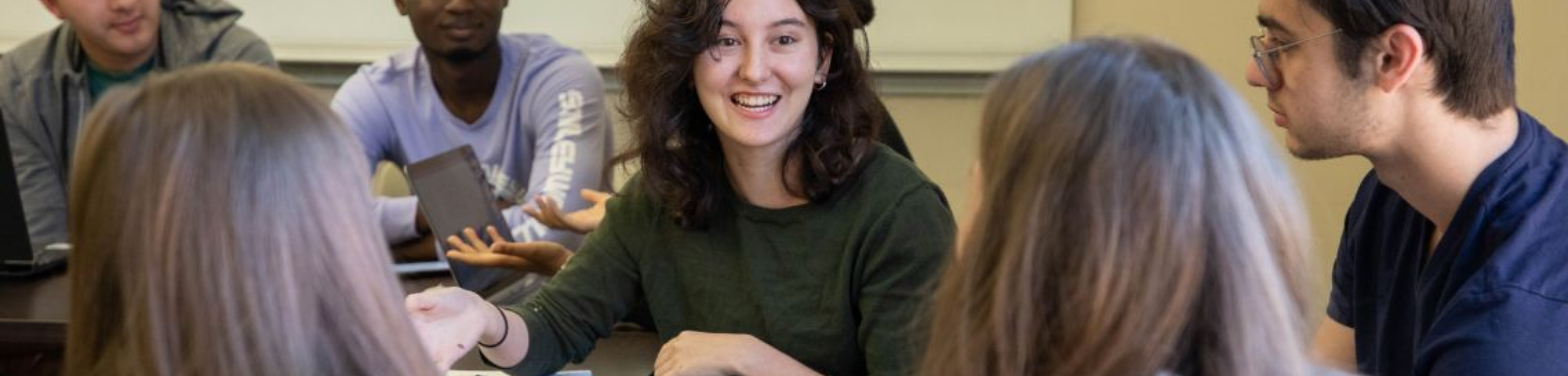 A group of students sit around a table in discussion, with one student in the center smiling and speaking while others listen.