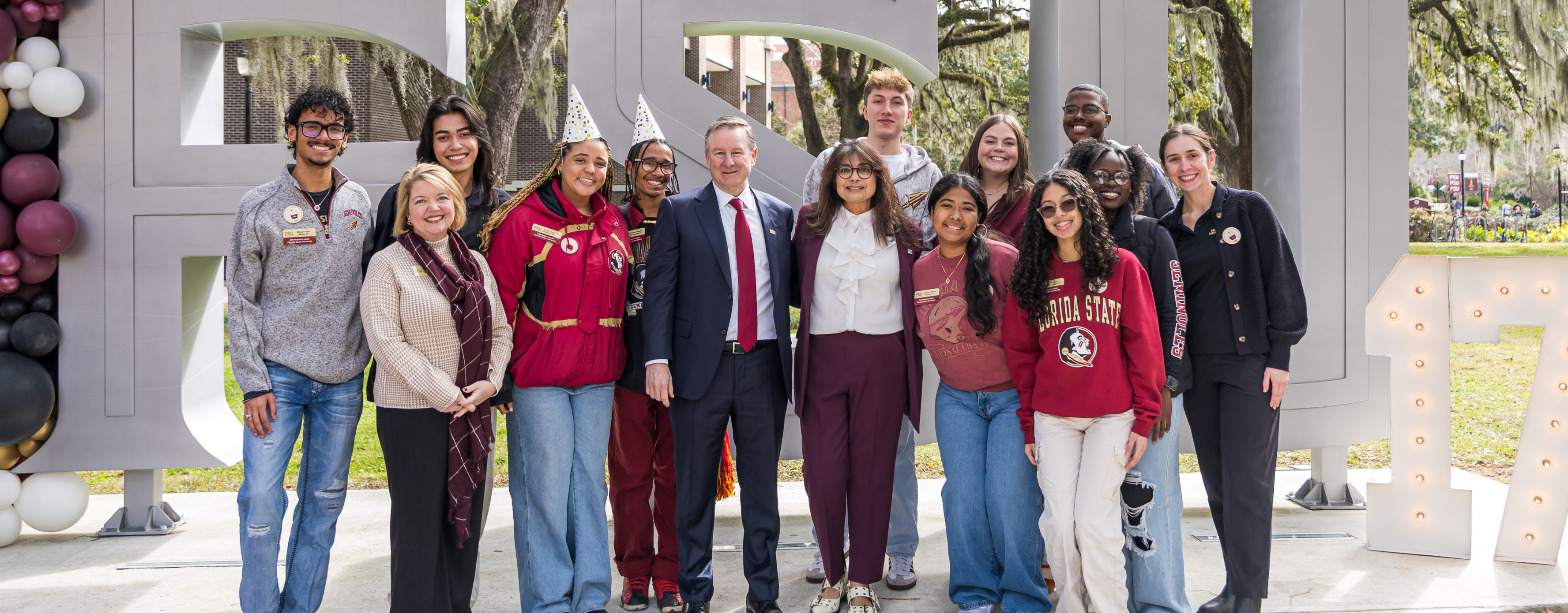 A group of about 15 students and staff members pose for a group photo in front of large, gray "FSU" block letters on the Florida State University campus.