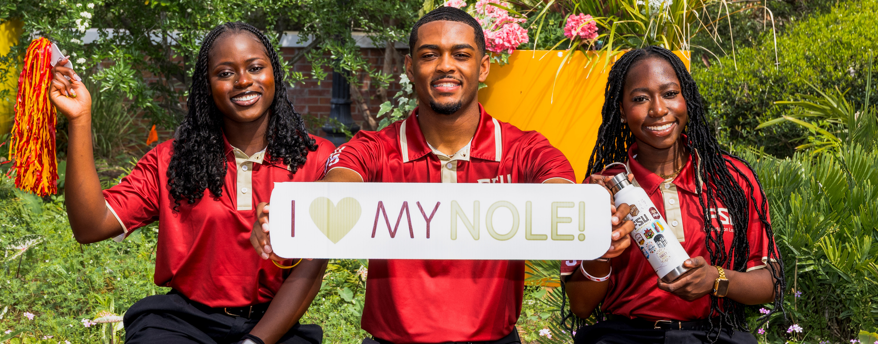 Three Florida State University students, two women and one man, are smiling and posing together outdoors in front of lush greenery. They are all wearing maroon FSU polo shirts with tan collars and trim. The man in the center holds a white sign that reads "I ❤️ MY NOLE!" in maroon and light green text. The woman on the left is holding a pom-pom with red and yellow streamers, and the woman on the right is holding an FSU-branded water bottle covered in stickers.