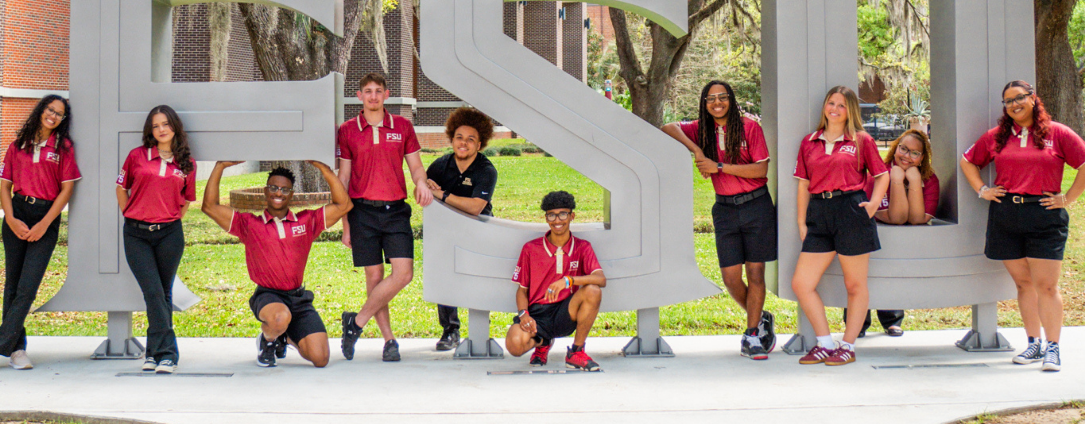A group of ten Florida State University student leaders are posing creatively around large, gray "FSU" block letters on campus.