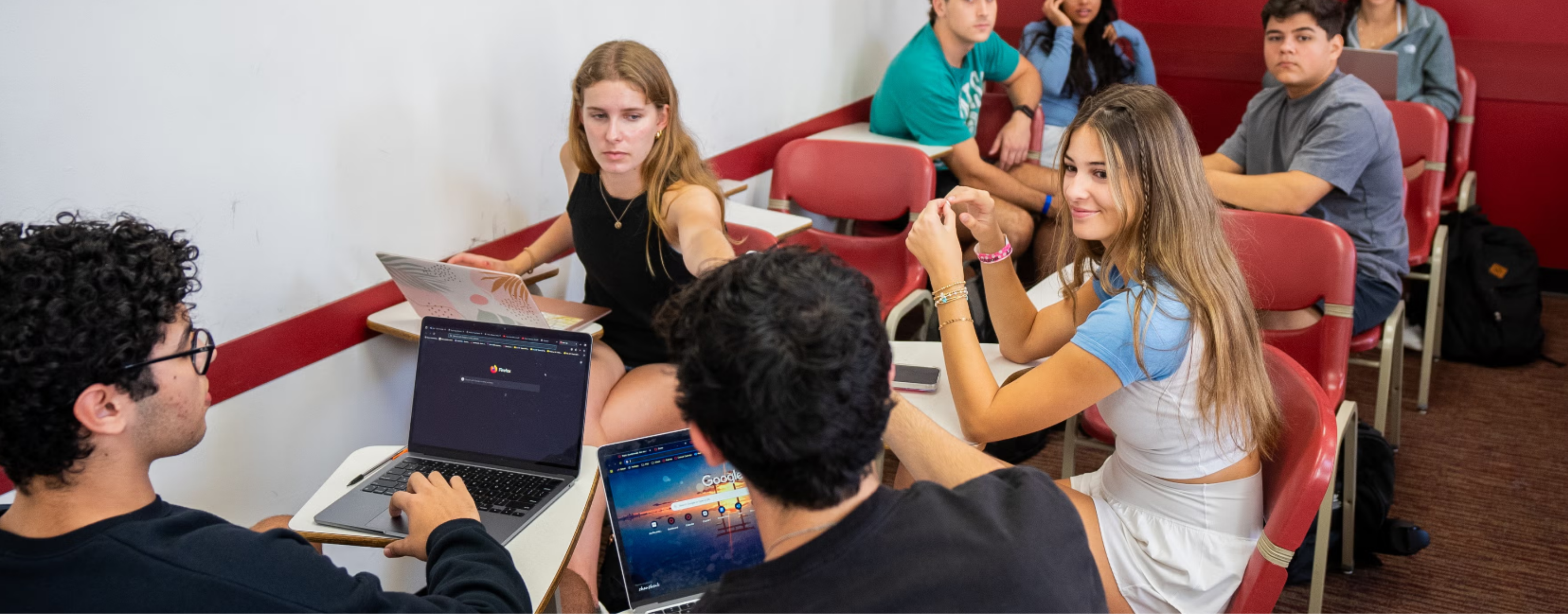 A group of students in a classroom are engaged in conversation, with some using laptops.