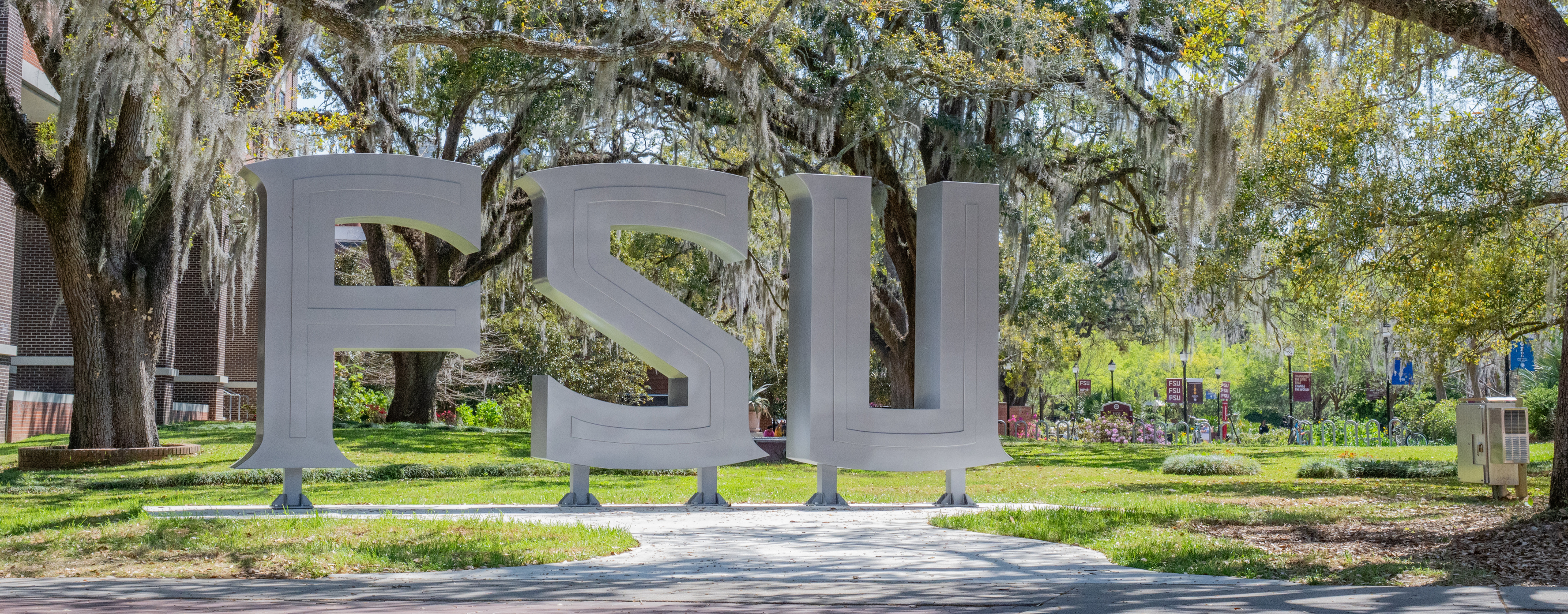 A large FSU sculpture stands prominently in a scenic, tree-filled campus setting.