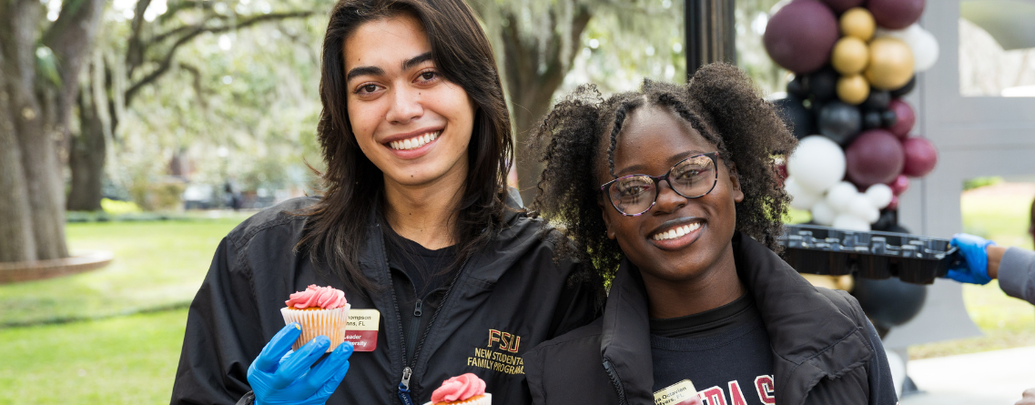 Two people smiling and holding cupcakes in an outdoor setting with balloons in the background.