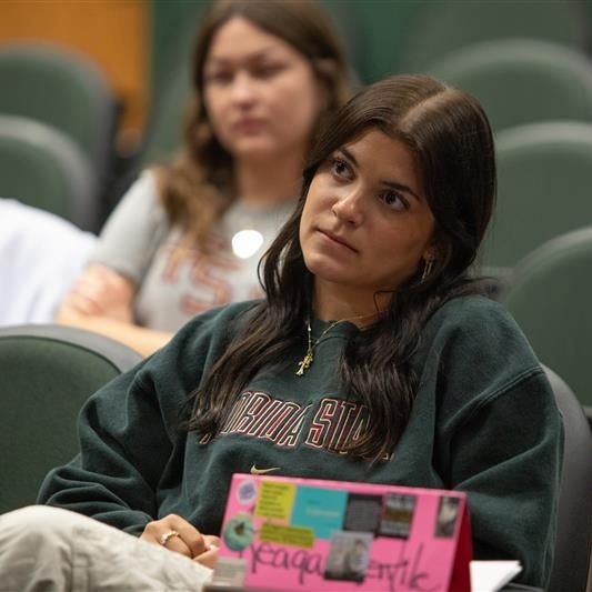 A young woman with long dark hair sits in a lecture hall or auditorium, looking forward with a focused expression. She is wearing a dark green sweater.